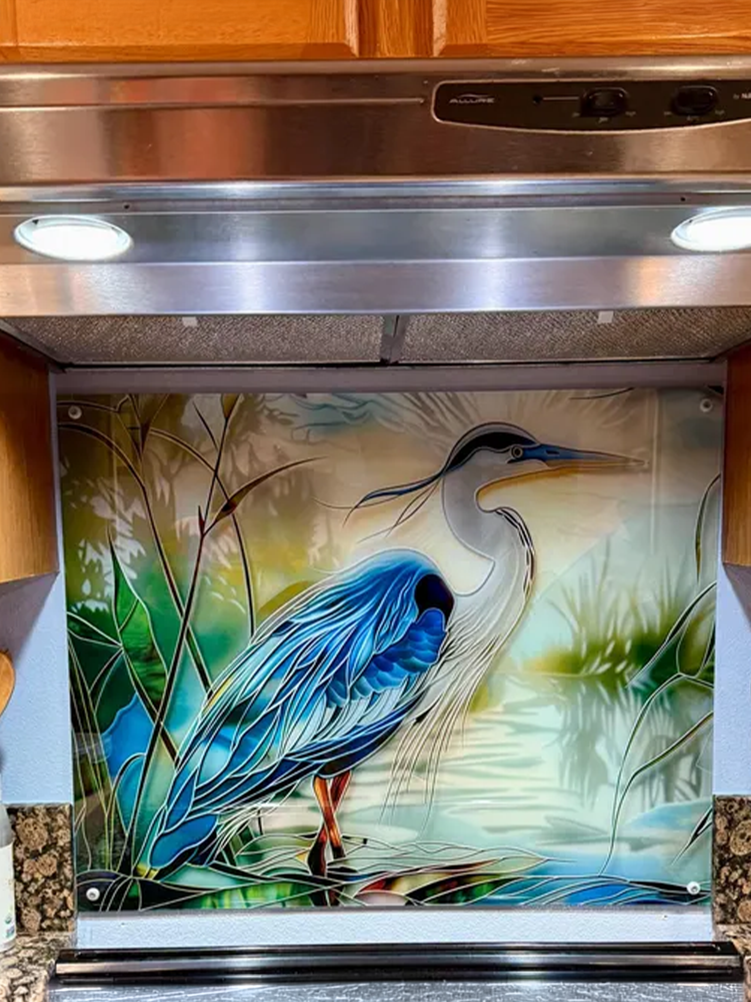 Tempered glass kitchen backsplash featuring a bright faux stained glass design of a majestic blue heron standing in a wetland, with bright sun rays radiating like a halo from behind its head, installed behind a modern stainless steel stove.