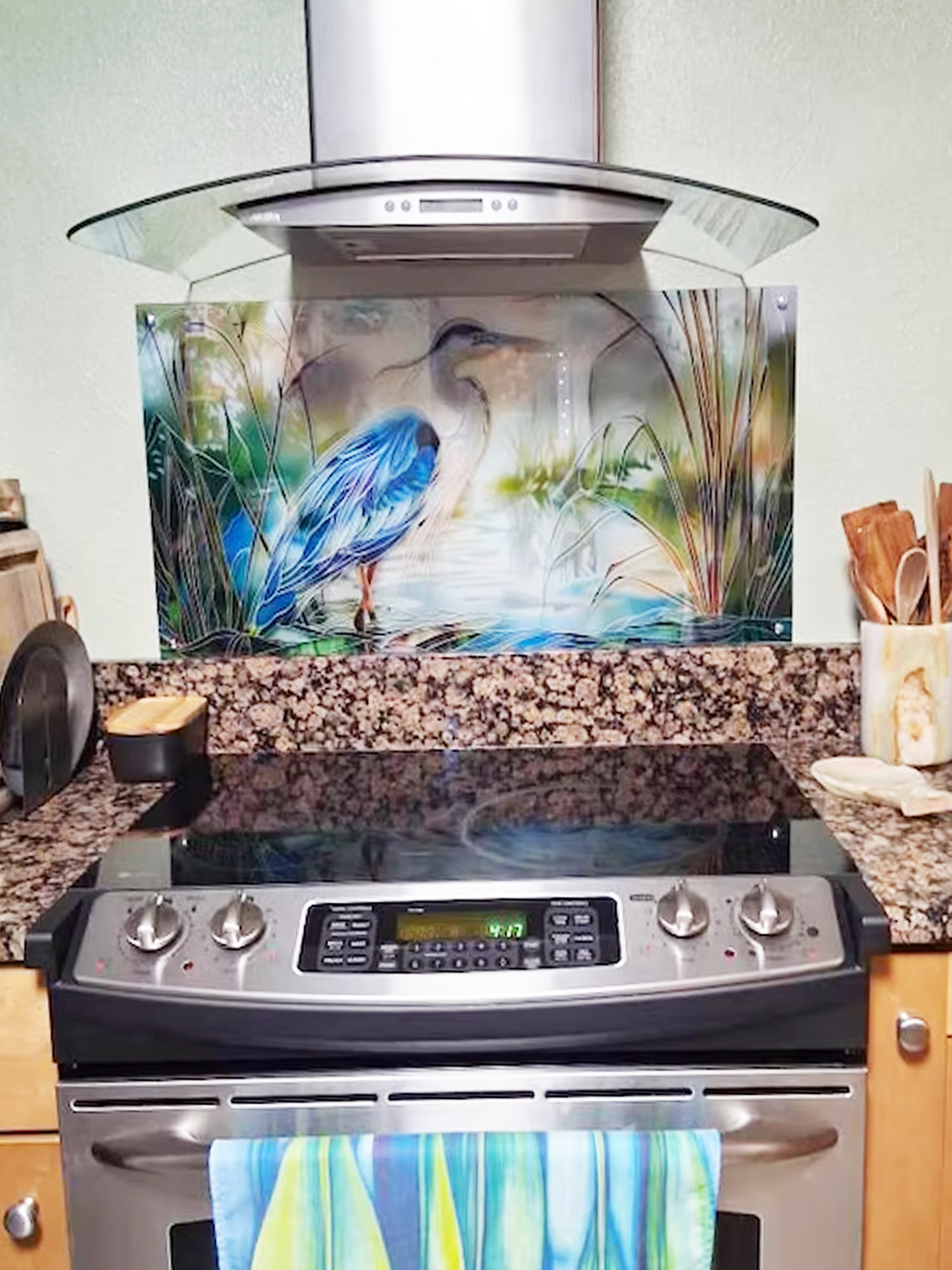 Tempered glass kitchen backsplash featuring a bright faux stained glass design of a majestic blue heron standing in a wetland, with bright sun rays radiating like a halo from behind its head, installed behind a modern stainless steel stove.