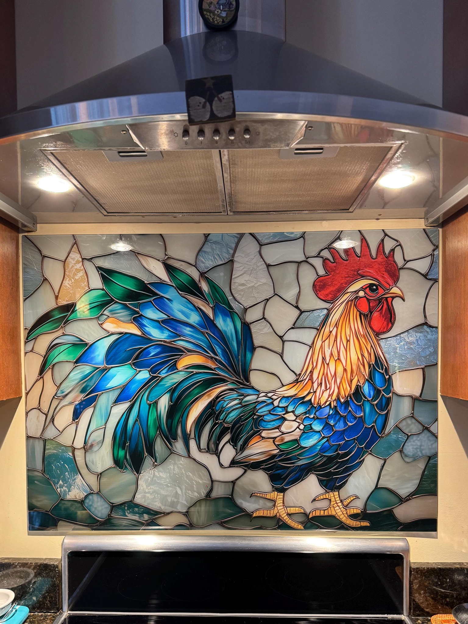 Tempered glass kitchen backsplash featuring a vibrant faux stained glass design of a rooster with striking teal and blue tail feathers against a light icy blue mosaic background, installed behind a modern stainless steel stove.