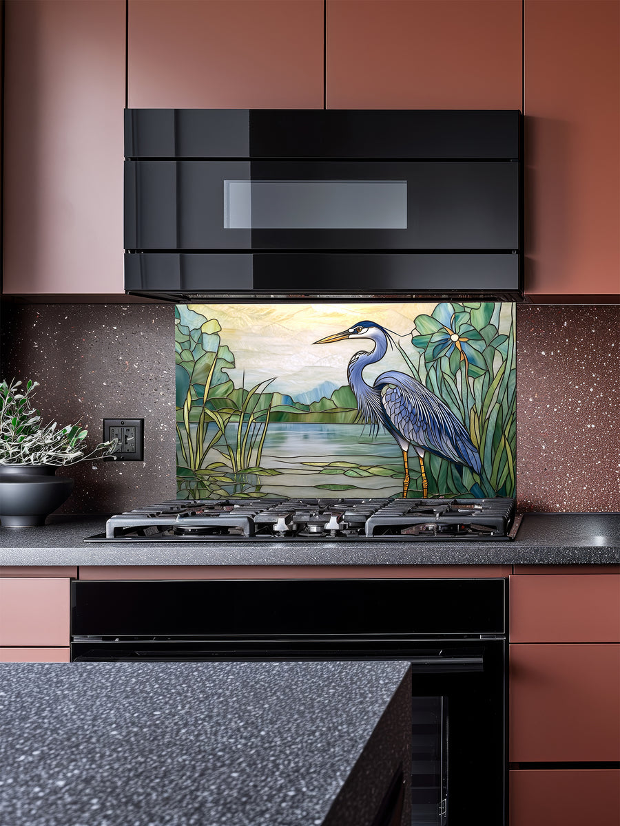 Tempered glass kitchen backsplash featuring a bright faux stained glass design of a majestic blue heron standing among lily pads against a glowing sunrise and mountain landscape, installed behind a modern stainless steel stove.